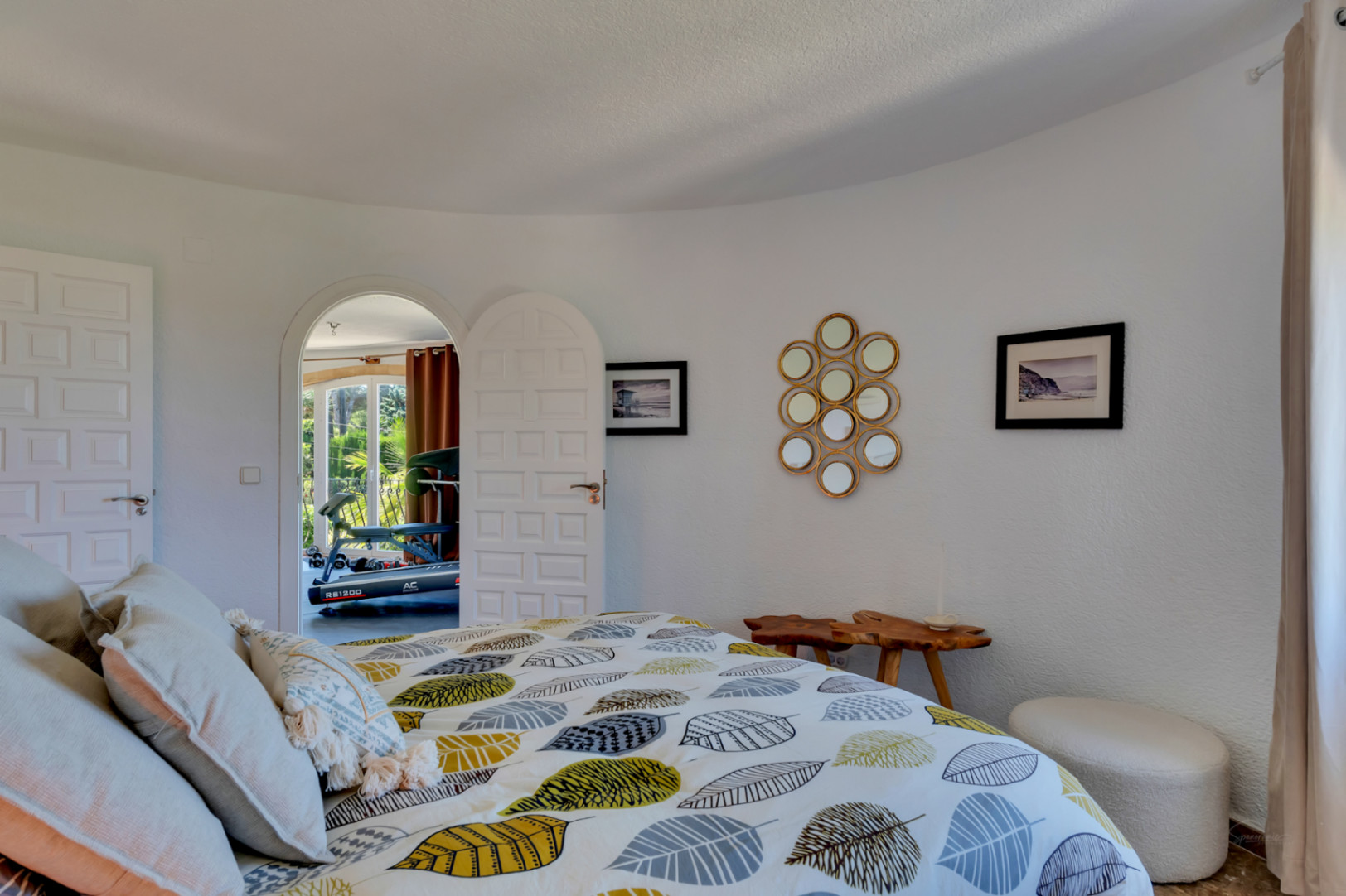 Dormitorio con paredes blancas texturizadas, cama de diseño con edredón de hojas y acceso arqueado a una sala luminosa con equipo de gimnasio.