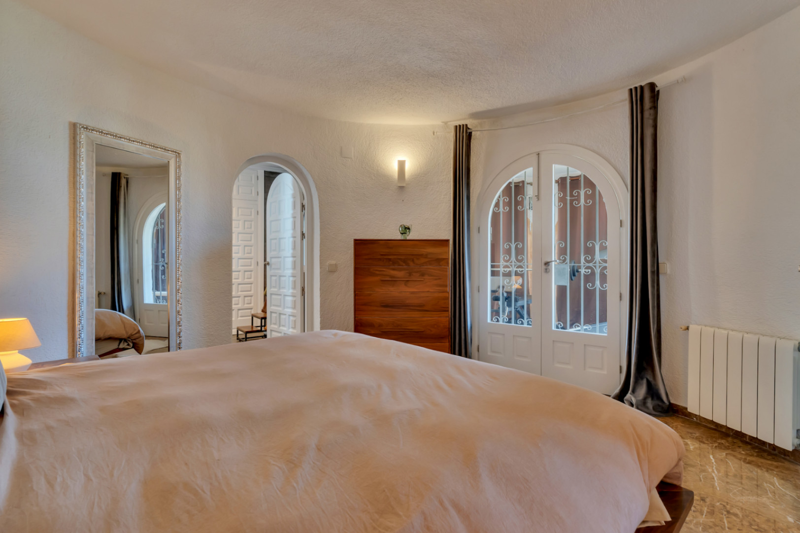 Dormitorio con puertas dobles arqueadas blancas y rejas al exterior, suelo de efecto mármol y cómoda de madera. Un gran espejo ornamental y paredes texturizadas completan la estancia.