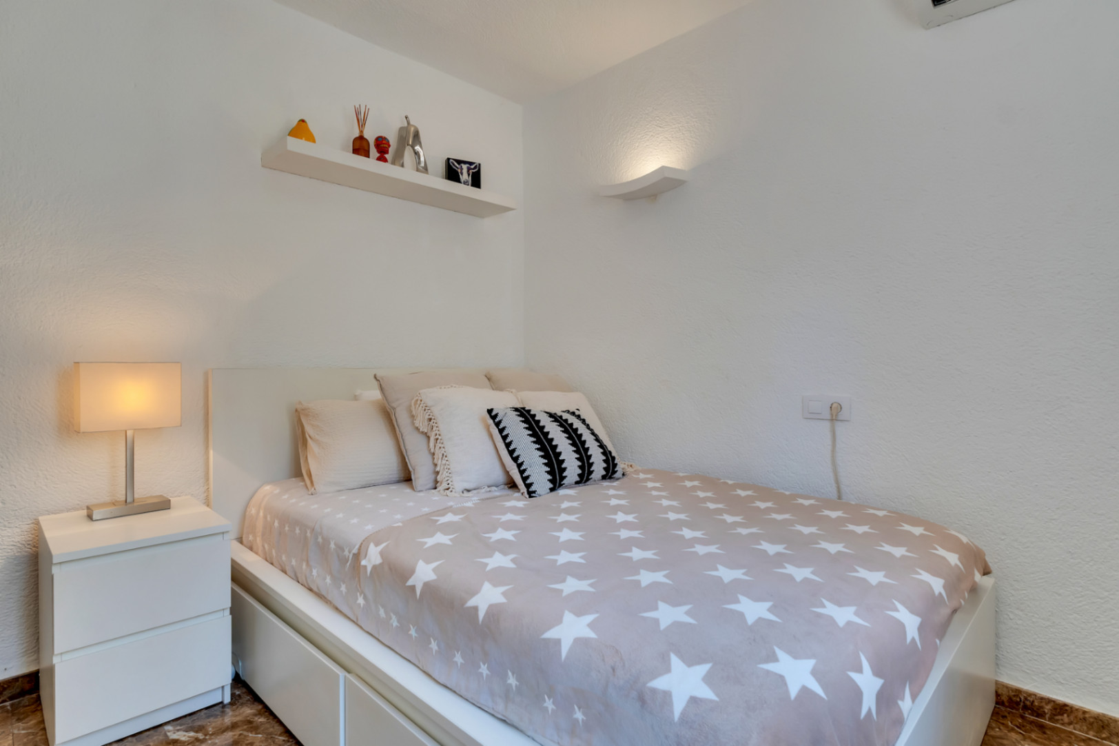 Dormitorio individual con cama moderna, couette de estrellas, mesita de noche blanca y estante decorativo. Iluminación mural y suelo de mármol.
