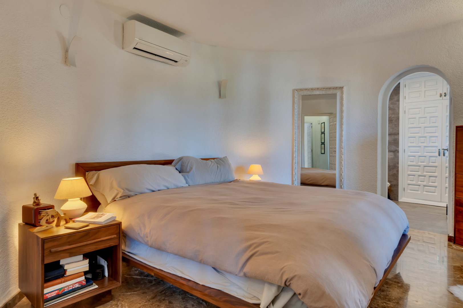 Dormitorio principal con cama de madera, paredes blancas texturizadas y aire acondicionado. Un espejo ornamentado y un arco conducen a una puerta de diseño.