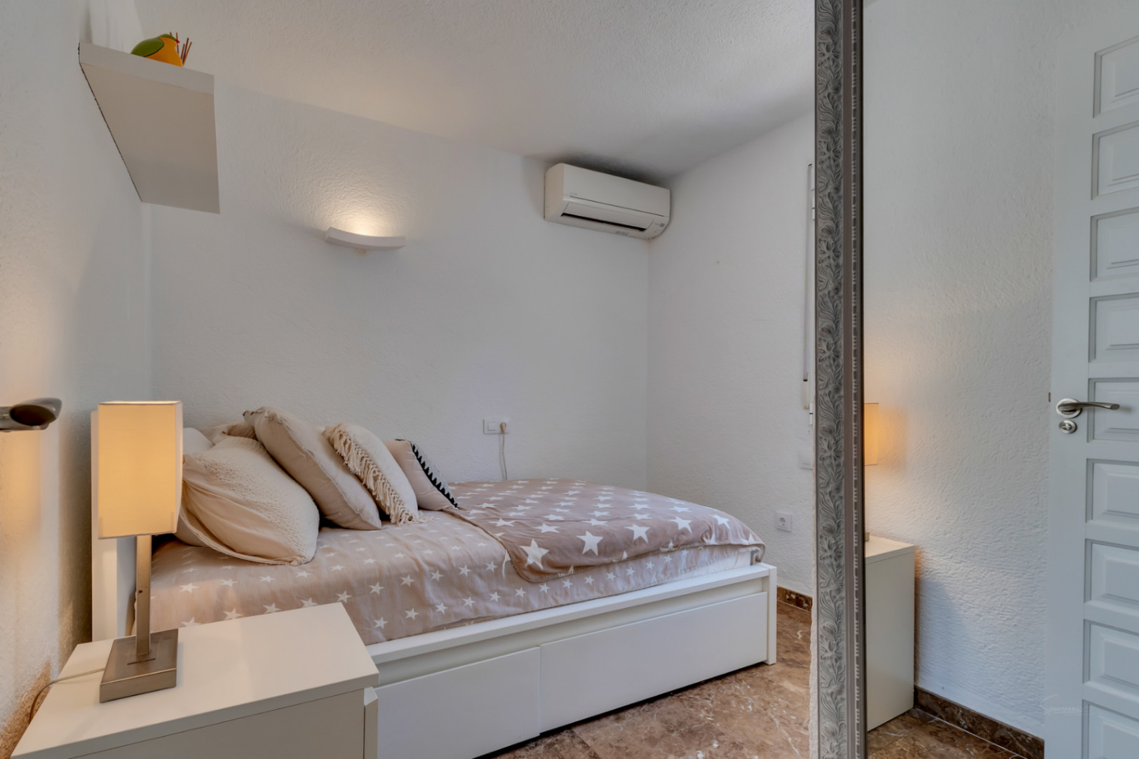Dormitorio secundario con paredes blancas texturizadas, cama con edredón de estrellas y mesita de noche con lámpara moderna. Incluye aire acondicionado y suelo de baldosas.