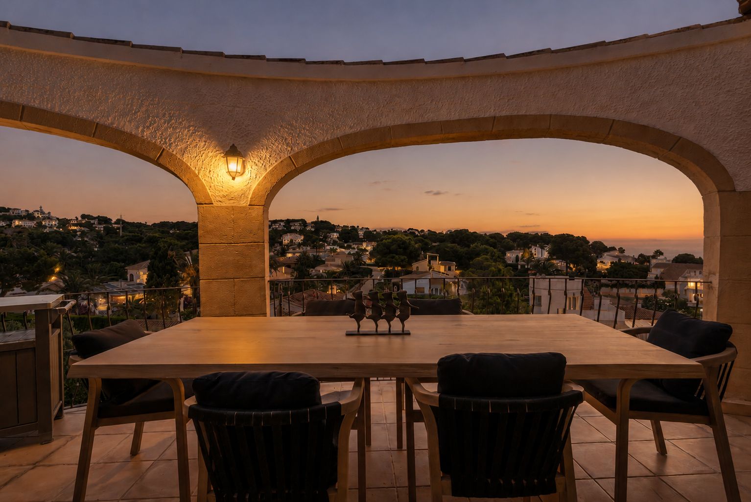 Elegante terraza con arcos al atardecer, equipada con una mesa de comedor de madera. Ofrece vistas panorámicas del área residencial iluminada y el horizonte marino.