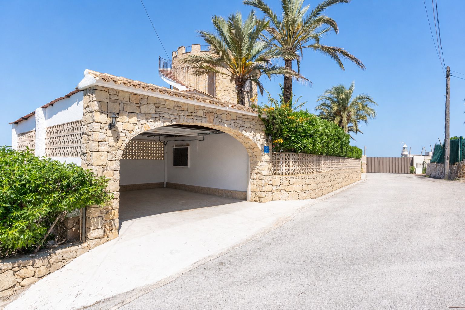 Entrada arqueada de piedra al garaje cubierto, integrada en la villa de estilo mediterráneo con escalera de caracol a la terraza superior. Palmeras y vegetación exuberante.