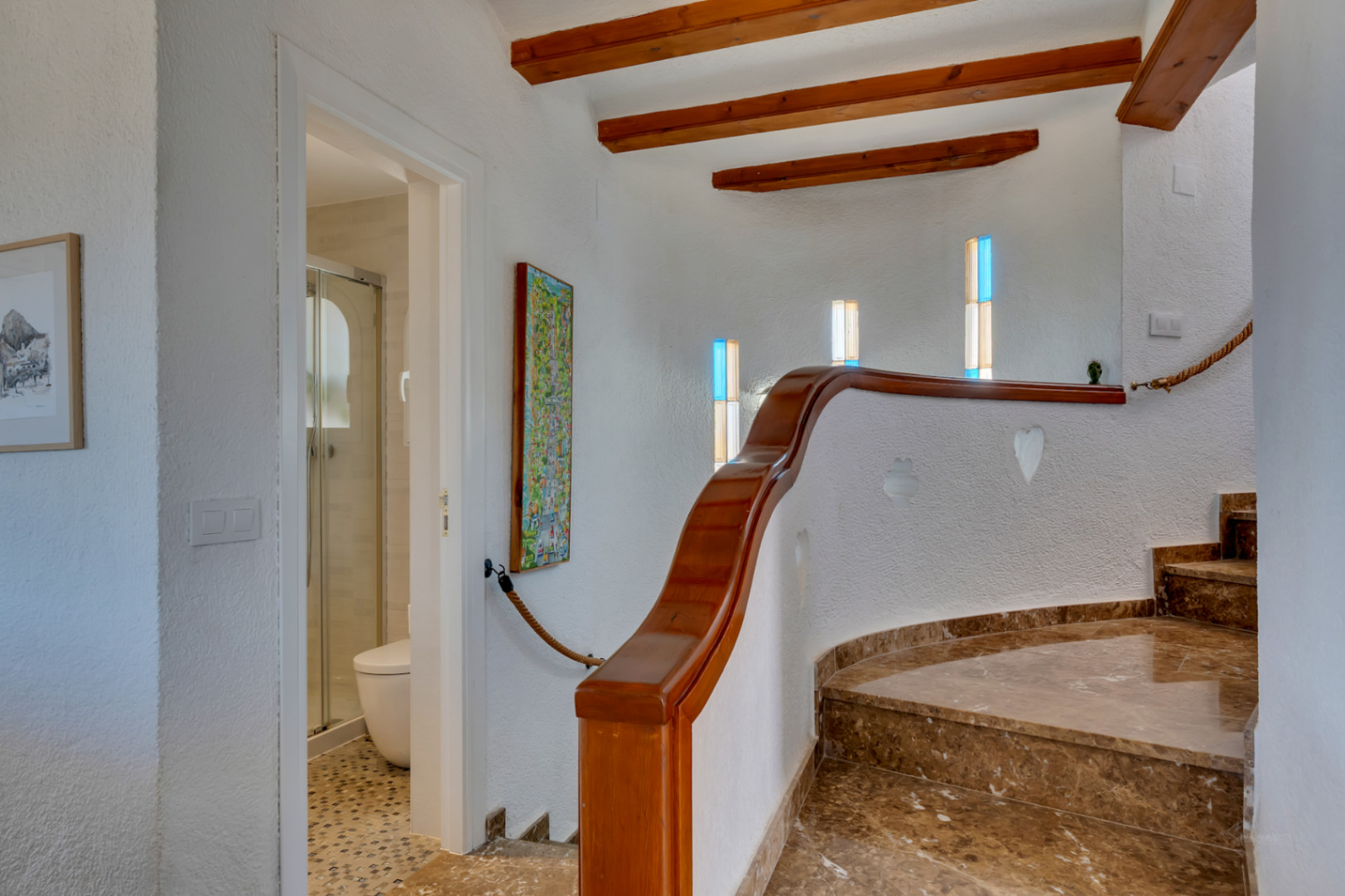 Escalera curva de mármol marrón con barandilla de madera, vigas expuestas y ventanas con vidrios de colores. Acceso a un baño con ducha.