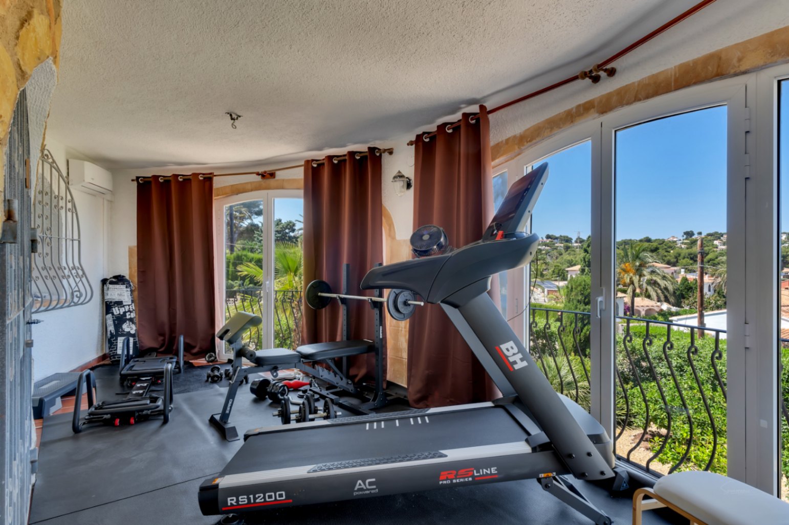Gimnasio en casa equipado con cinta de correr y pesas, ofreciendo vistas panorámicas a la vegetación y al paisaje desde sus grandes ventanales.