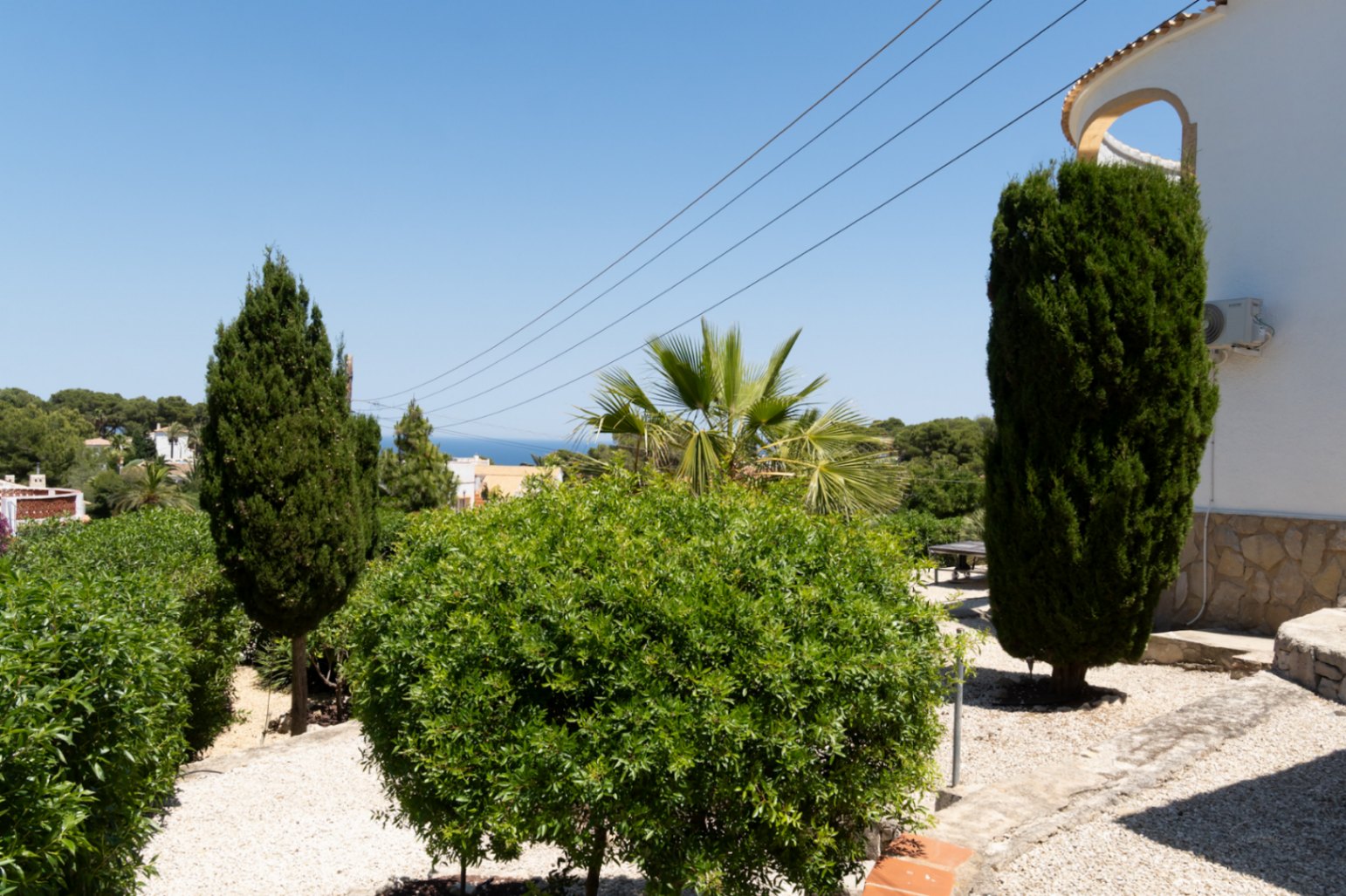 Jardín mediterráneo con sendero de grava, exuberante vegetación y cipreses, ofreciendo una vista lejana al mar bajo un cielo azul.