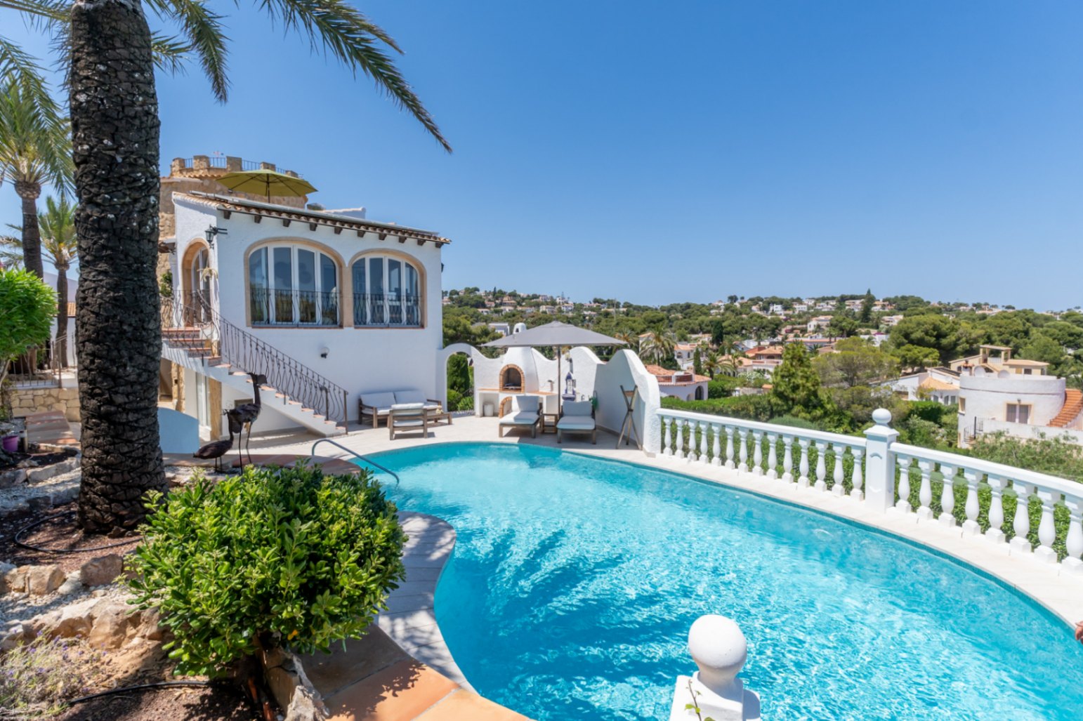 La piscina curva de agua azul cristalina se integra con la villa mediterránea blanca, que presenta ventanas arqueadas y una zona de barbacoa exterior con tumbonas, en un entorno verde.
