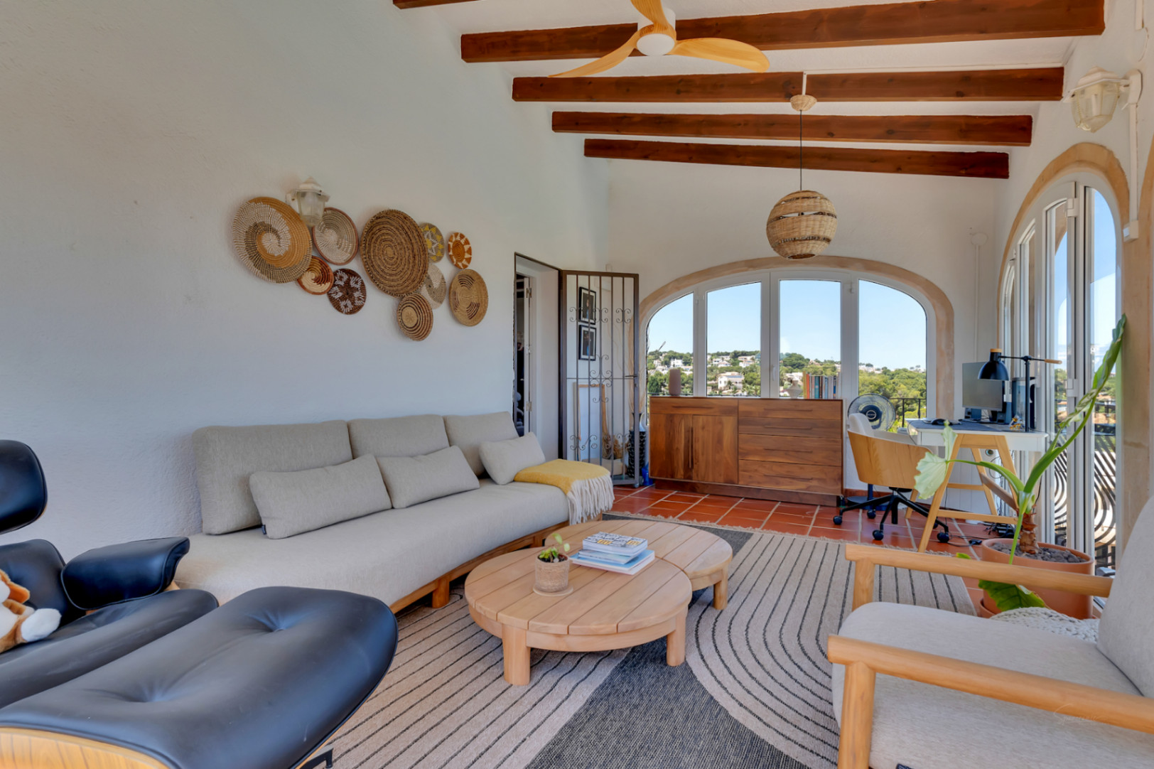 Luminosa terraza acristalada con vigas de madera vistas, suelos de terracota y ventanas arqueadas que ofrecen vistas al paisaje. Integra zona de estar y escritorio.