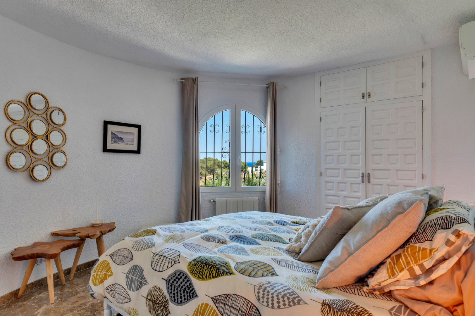 Luminoso dormitorio con ventana arqueada y rejas de hierro forjado, que ofrece vistas parciales al mar. Cuenta con armarios empotrados blancos y mesas auxiliares de madera rústica.