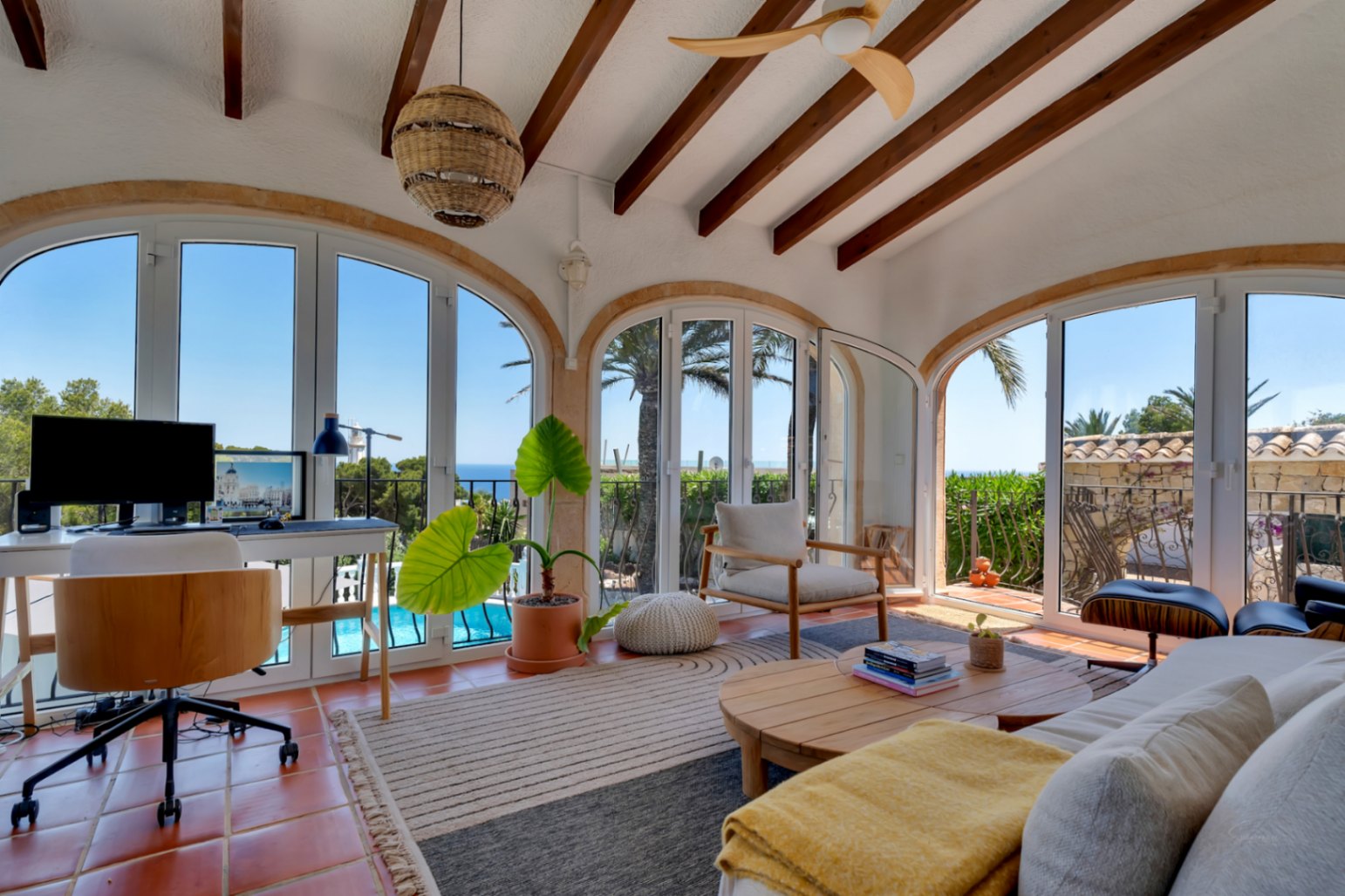 Luminoso salón con suelo de terracota y vigas de madera, que ofrece un espacio de trabajo con vistas panorámicas al mar y la piscina a través de sus ventanales arqueados.