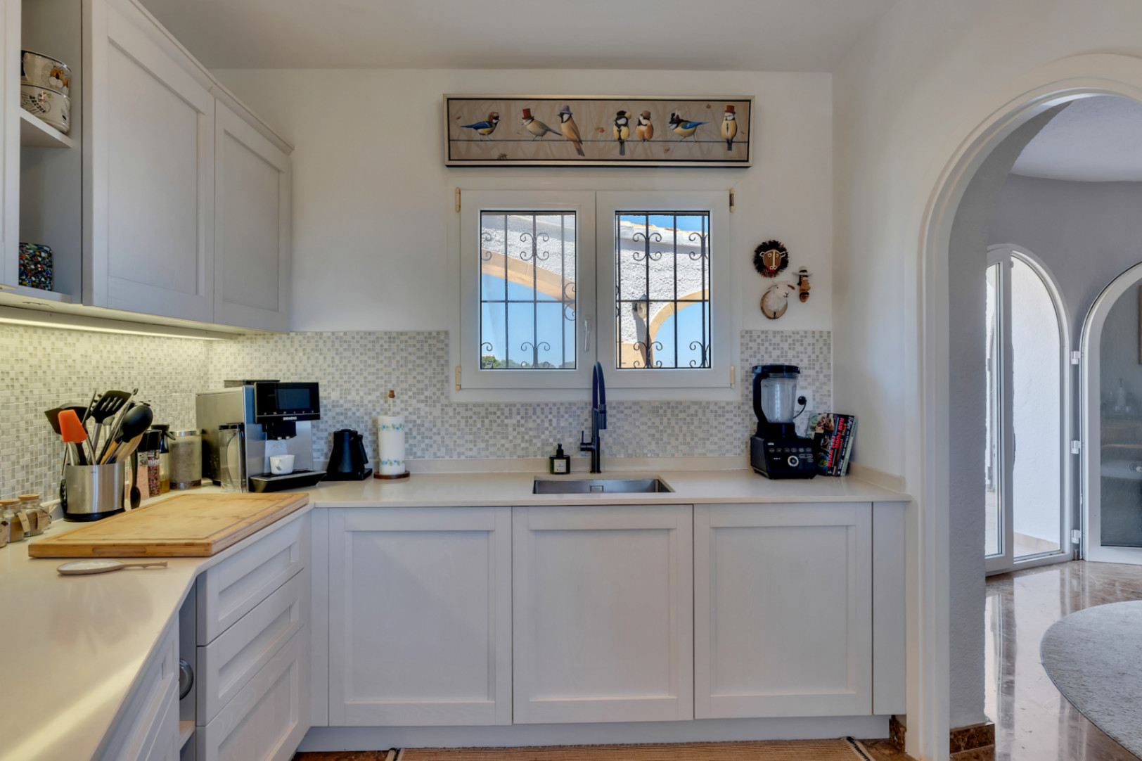 Moderna cocina con armarios blancos, encimeras claras y salpicadero de mosaico. Una ventana con rejas ofrece vistas al exterior, y un arco conduce a un espacio adyacente luminoso.
