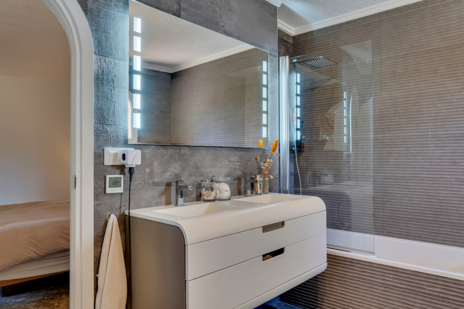 Moderno baño con doble lavabo y mueble curvo, espejo iluminado con LED y paredes de azulejos texturizados en gris oscuro. La ducha integrada con mampara de cristal complementa el diseño.