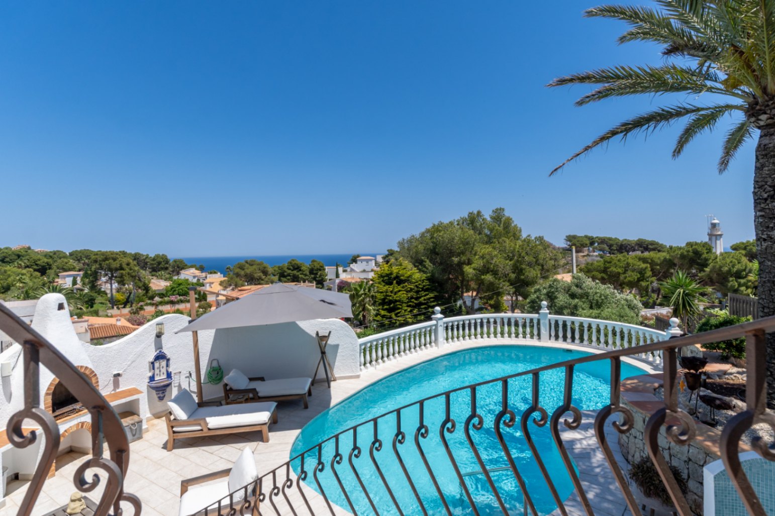 Piscina curva con terraza de azulejos, zona de barbacoa tradicional y vistas panorámicas al mar Mediterráneo y al faro.