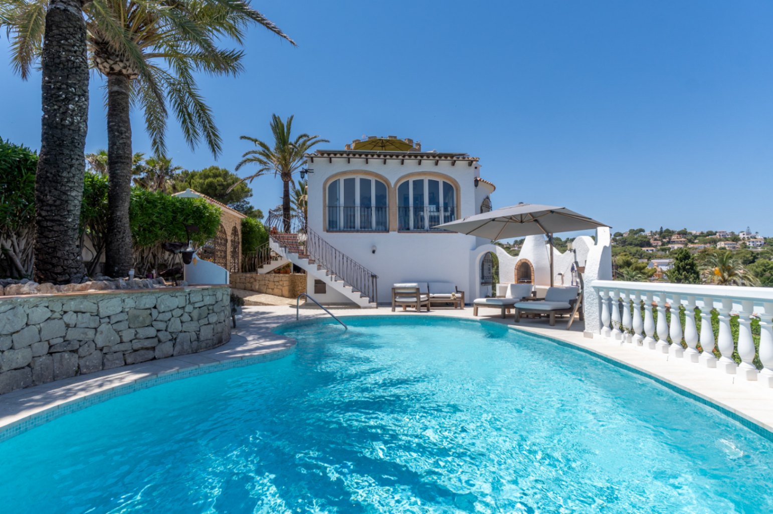 Piscina curva de agua azul con zona de solárium, barbacoa exterior y una villa mediterránea de fachada blanca, rodeada de palmeras y vegetación.