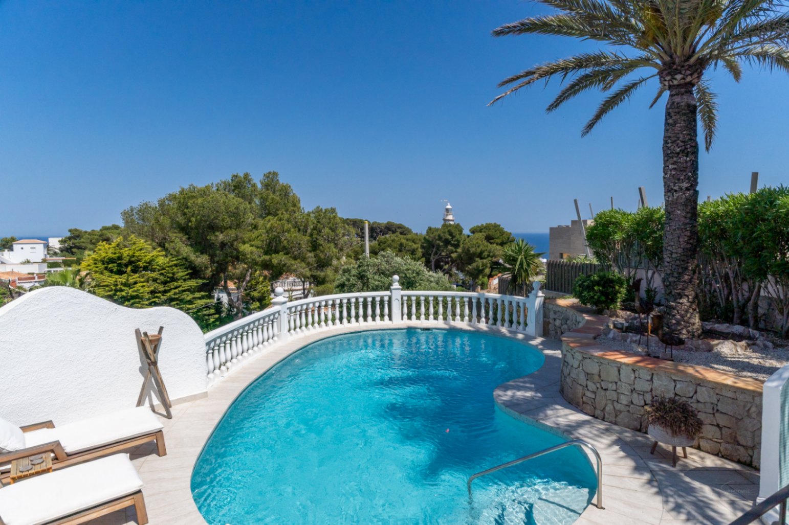 Piscina de diseño curvo con balaustrada blanca, tumbonas y palmera. Ofrece vistas lejanas al mar y un faro bajo un cielo despejado.
