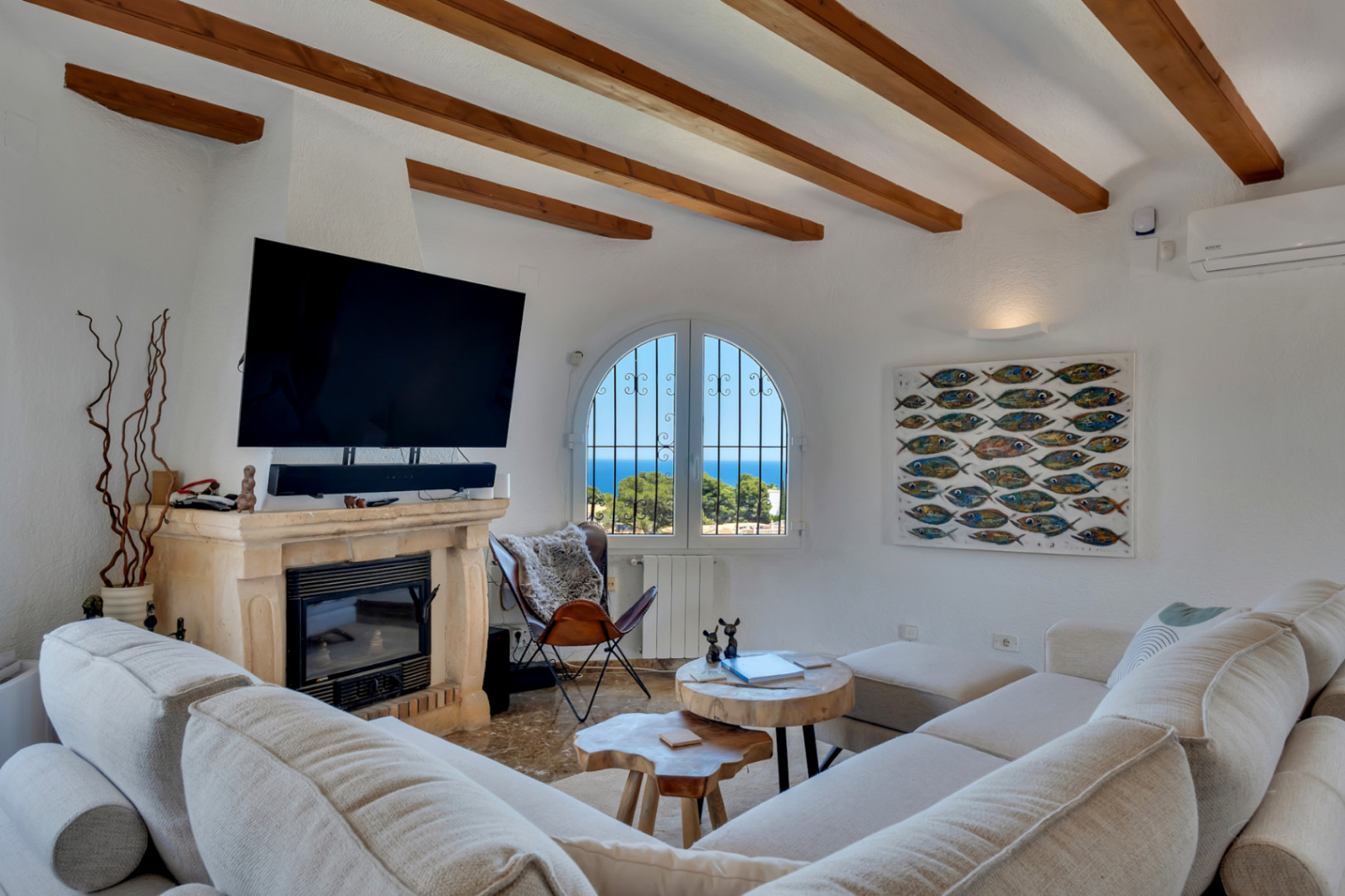 Salón con vigas de madera a la vista y chimenea de piedra, ofreciendo vistas al mar a través de una ventana arqueada. Sofá modular y mesas de centro de madera natural.