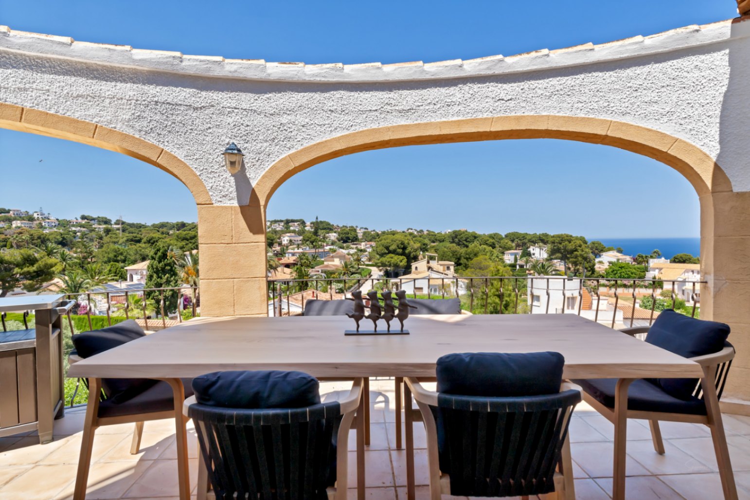 Terraza cubierta con mesa de comedor de madera clara y sillas con cojines oscuros, ofreciendo vistas panorámicas al mar y al paisaje urbano a través de arcos mediterráneos.