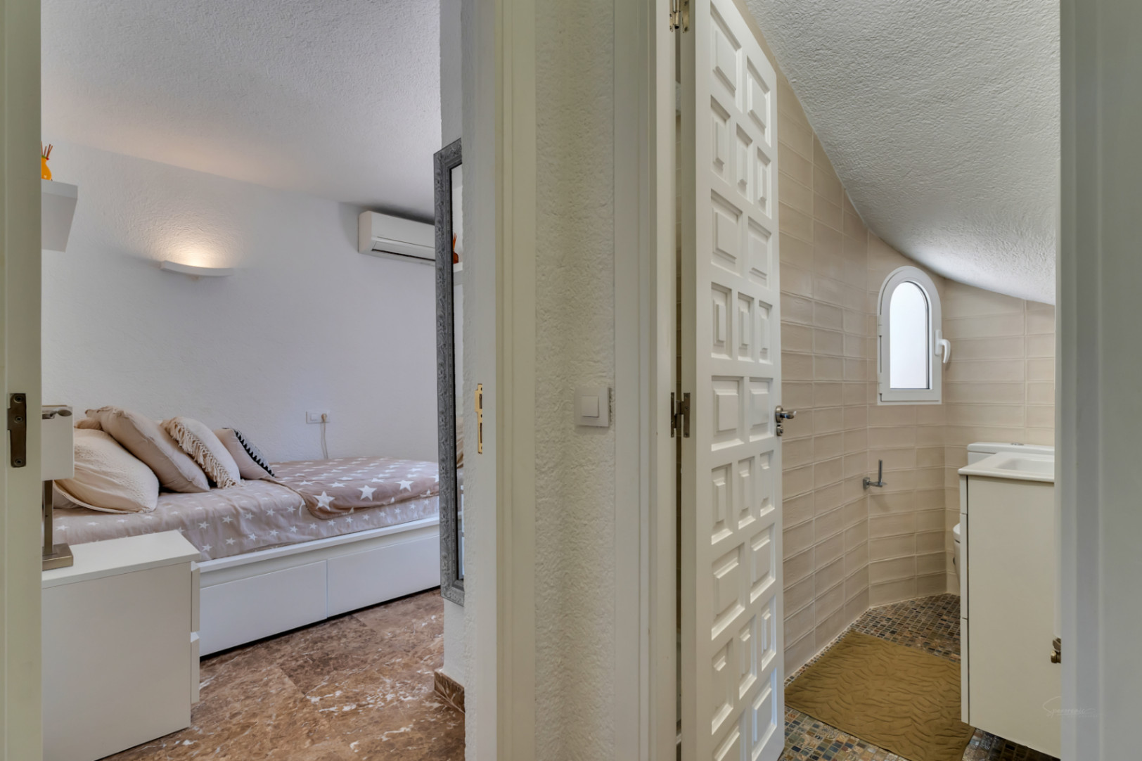 Vista a un dormitorio con suelo efecto mármol y aire acondicionado, frente a un baño con paredes alicatadas en beige, ventana arqueada y suelo de mosaico con lavabo blanco.