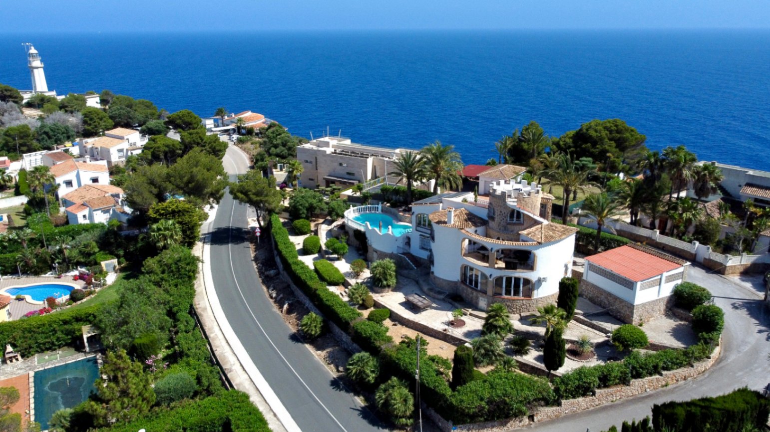 Vista aérea de la villa mediterránea en Jávea, con piscina elevada, jardines aterrazados y arquitectura tradicional, frente al vasto mar azul y un faro lejano.