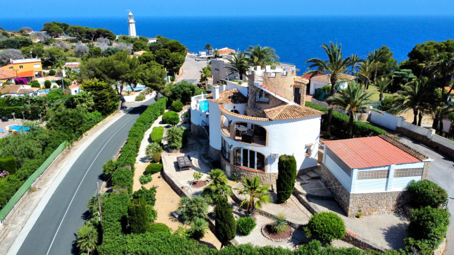 Vista aérea de una villa mediterránea con paredes blancas, tejado de tejas y piscina. Jardines exuberantes rodean la propiedad, con el mar y un faro al fondo.