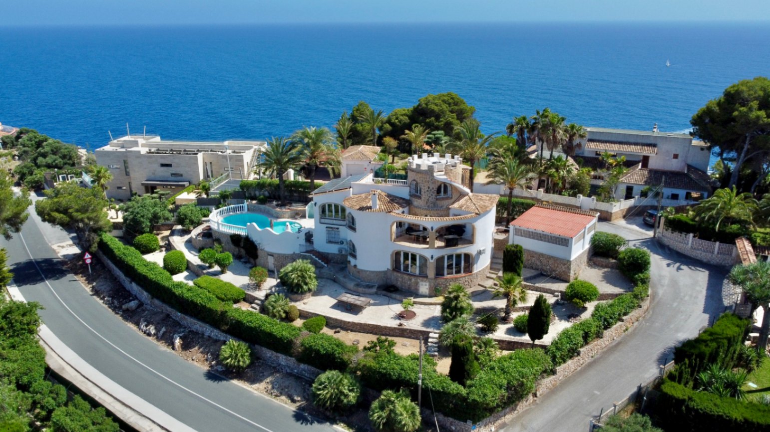 Vista aérea de una villa mediterránea con torre circular, jardines escalonados y piscina de forma libre, todo con el mar azul de fondo.