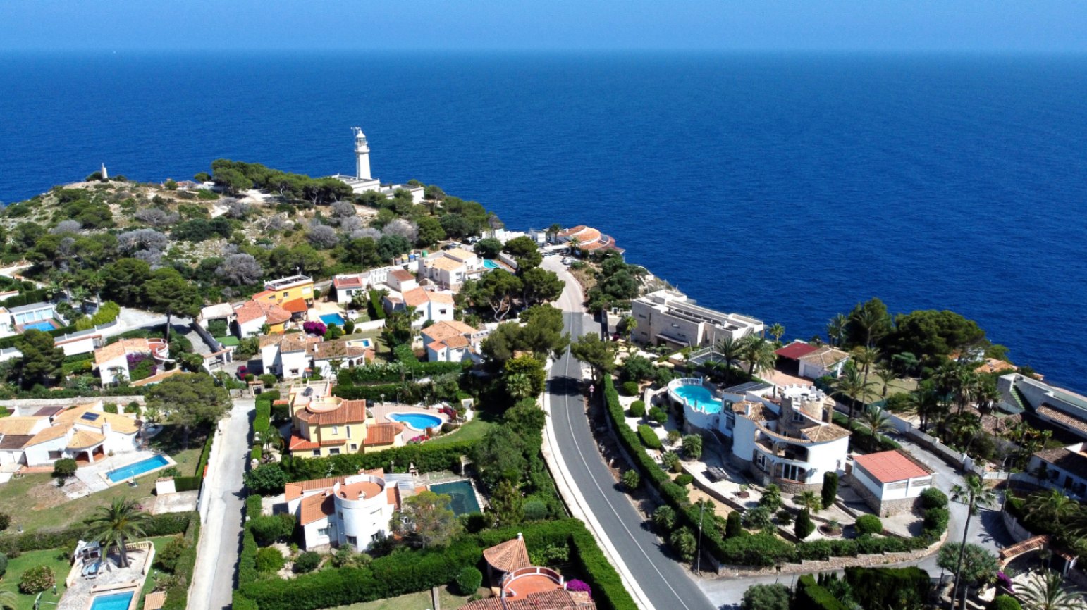 Vista aérea panorámica de una urbanización costera con villas, piscinas privadas y jardines exuberantes, bordeando el azul profundo del Mediterráneo y un faro lejano.