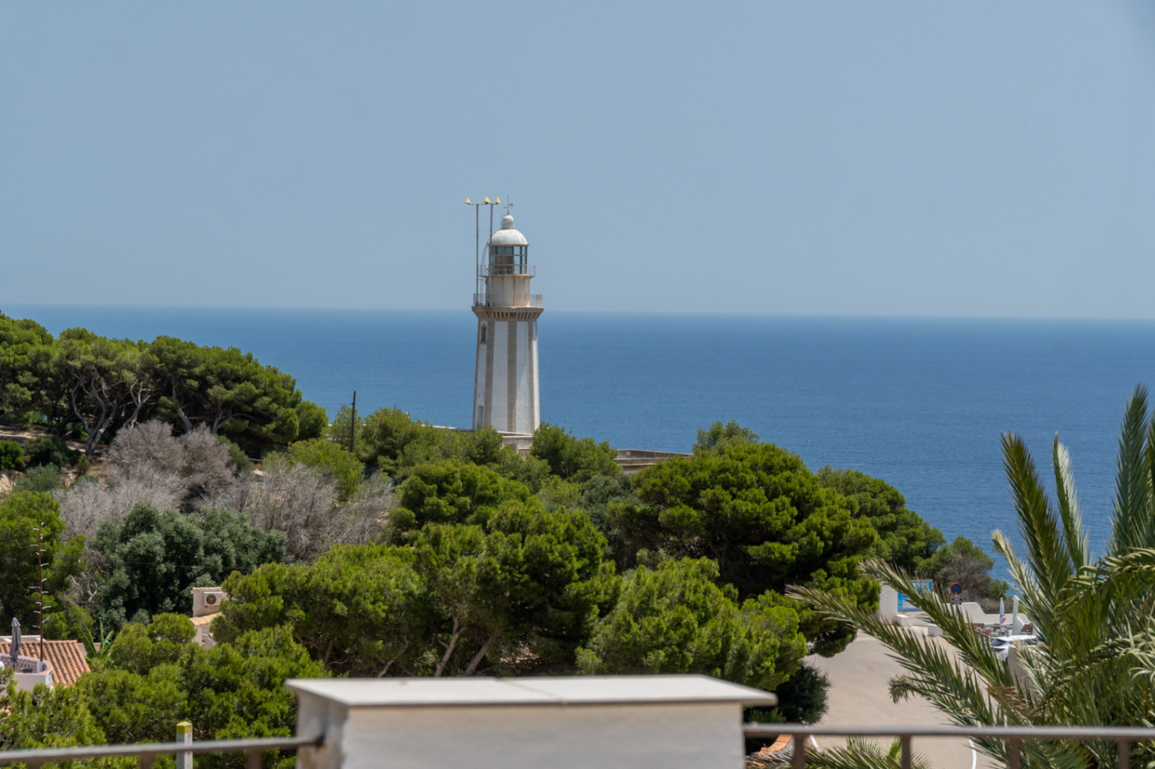 Vista del faro blanco con rayas grises sobre una colina verde, enmarcado por el Mediterráneo azul y un cielo despejado, con vegetación exuberante en primer plano.