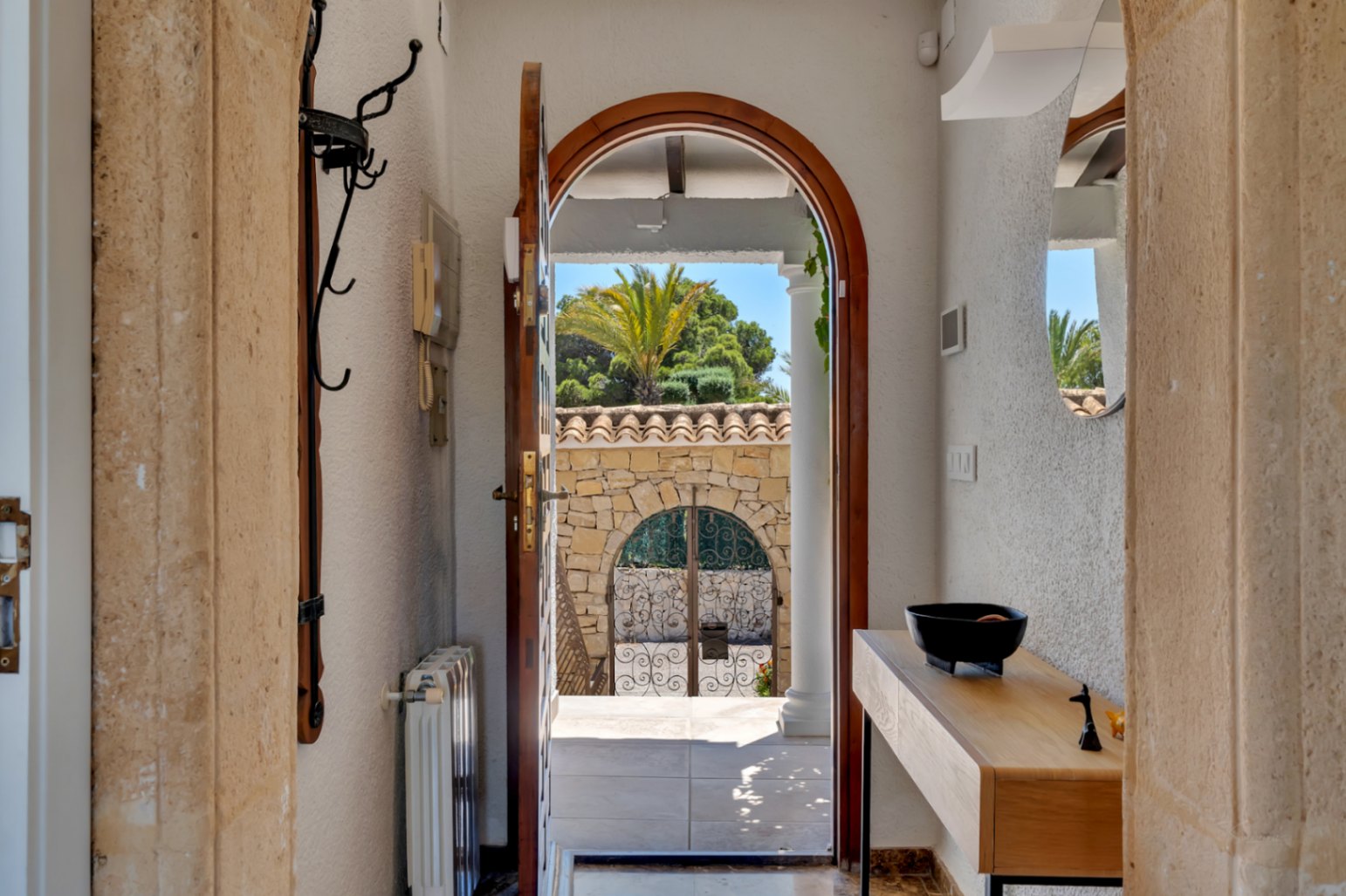 Vista desde el recibidor a través de una puerta de madera arqueada hacia un muro de piedra con verja de hierro forjado y exuberante vegetación mediterránea. En el interior, consola de madera y espejo.