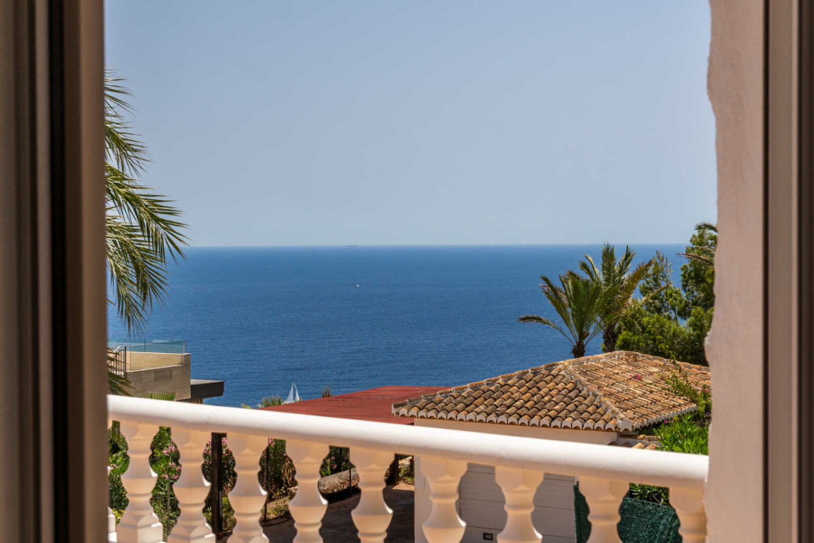 Vista desde un balcón con balaustrada blanca, tejado de terracota y palmeras, todo con vistas al vasto mar Mediterráneo y un velero en la distancia.