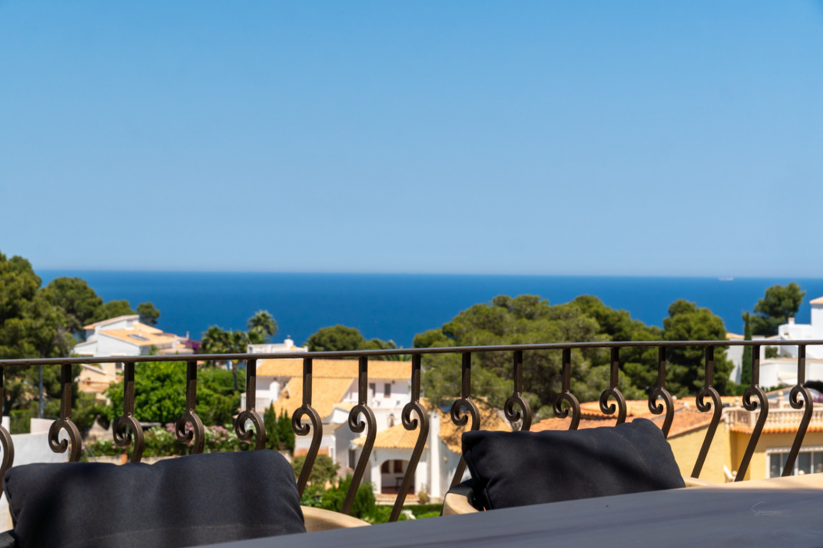 Vista desde una terraza con barandilla de hierro forjado, mostrando tejados de terracota entre pinos y el mar Mediterráneo.