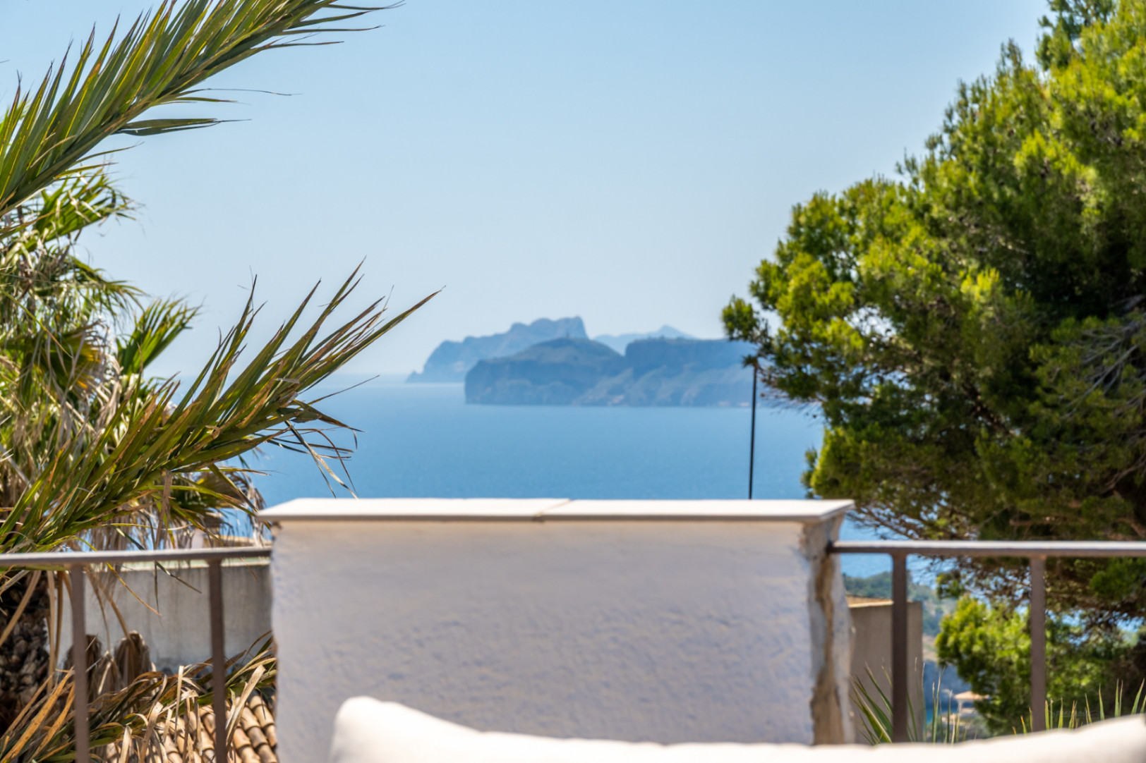 Vista panorámica al mar con una isla montañosa en el horizonte, enmarcada por vegetación mediterránea y una barandilla de terraza.