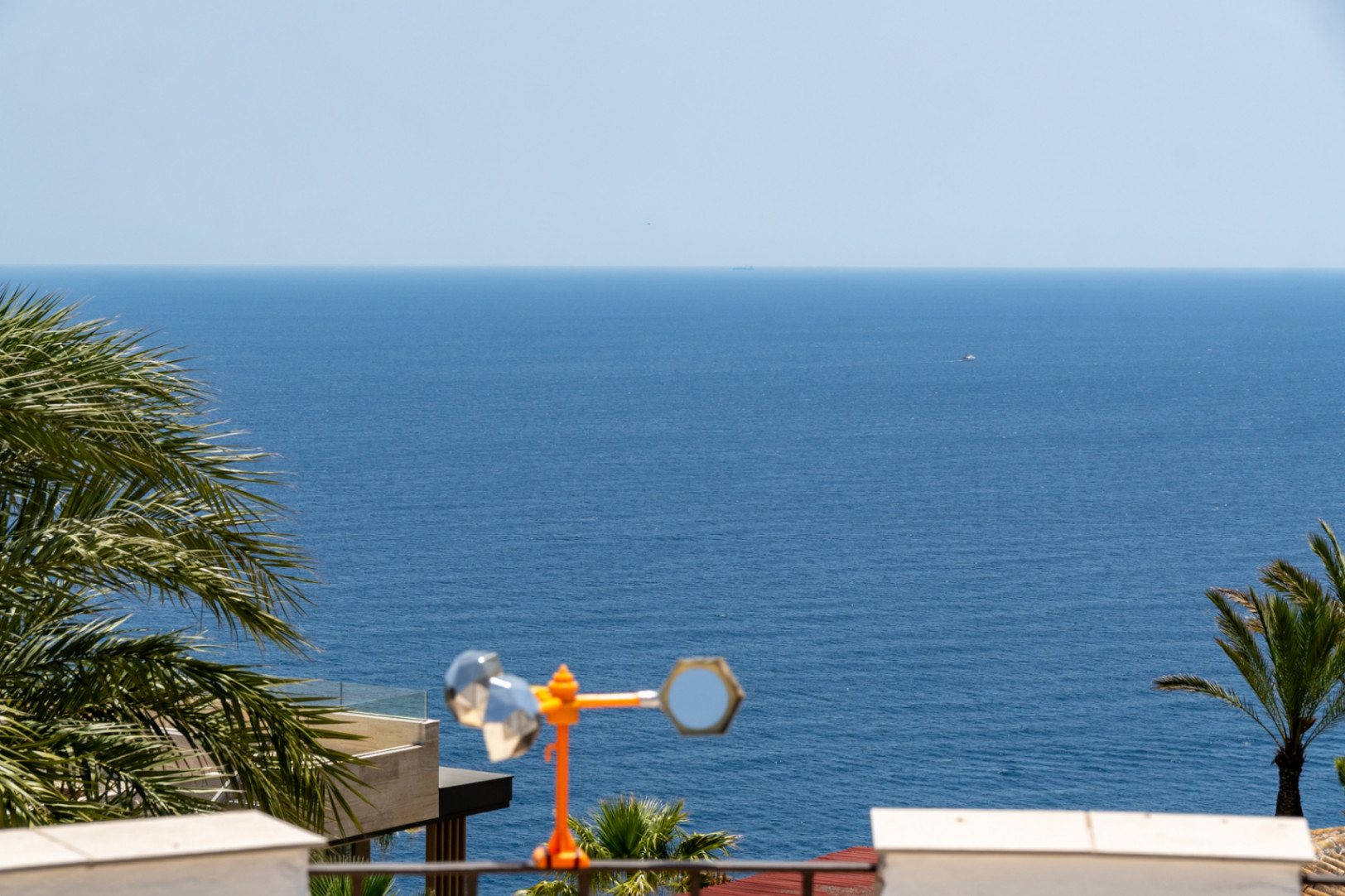 Vista panorámica del mar Mediterráneo enmarcada por palmeras, con el borde de una terraza moderna y un anemómetro decorativo en primer plano.