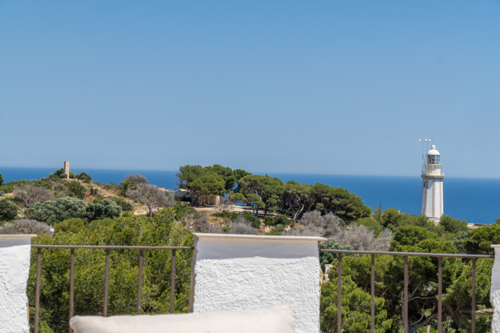 Vista panorámica desde la terraza con barandilla de metal, mostrando el mar Mediterráneo, el faro y una torre de piedra en la distancia.