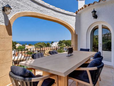 Amplia terraza comedor con mesa de madera clara y sillas con cojines azules. Un arco enmarca vistas panorámicas al mar Mediterráneo y la costa.