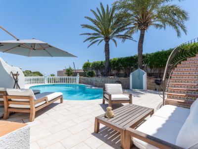 Amplia terraza junto a la piscina con tumbonas de madera, un conjunto de sofás exterior y una escalera curva con azulejos decorativos. Palmeras y una ducha exterior complementan el espacio.