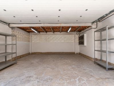 Amplio garaje con suelo de hormigón texturizado, paredes de estuco blanco y bloques decorativos de ventilación. Incluye tejas de terracota vistas, iluminación fluorescente y estanterías metálicas.