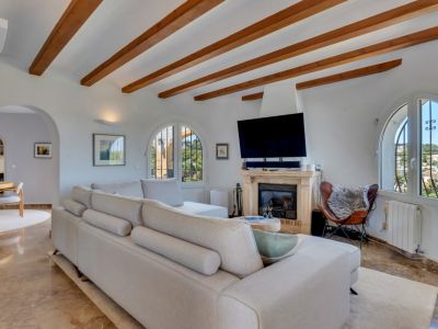 Amplio salón con techo de vigas de madera, chimenea de piedra y ventanales arqueados que ofrecen vistas al exterior. Sofá modular claro y suelos de mármol.