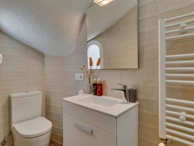 Aseo funcional con paredes de azulejos claros, mueble de lavabo blanco y un inodoro. Un espejo refleja una ventana arqueada y un toallero calefactable.