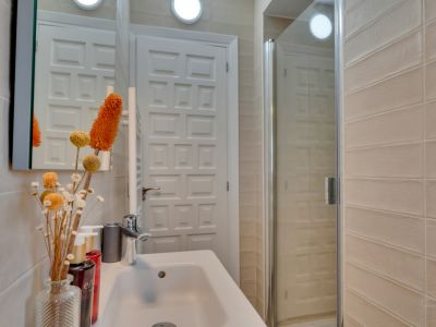 Baño compacto con lavabo blanco, grifería cromada y ducha con mampara de cristal. Destacan los azulejos beige y la puerta panelada.