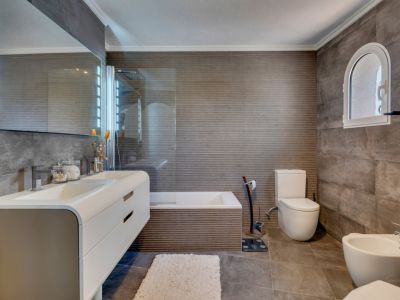 Baño moderno con azulejos grises texturizados, un mueble de doble lavabo flotante y un espejo iluminado. Una ventana arqueada añade carácter sobre la zona del inodoro.