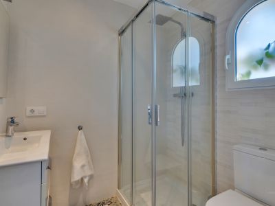 Baño moderno con cabina de ducha de cristal y rociador de lluvia. Destaca una ventana arqueada esmerilada y un lavabo con mueble blanco, junto a azulejos texturizados.