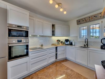 Cocina equipada con armarios blancos estilo shaker, electrodomésticos integrados de acero inoxidable, encimera clara y salpicadero de mosaico. Ventana con rejas decorativas y vistas verdes.