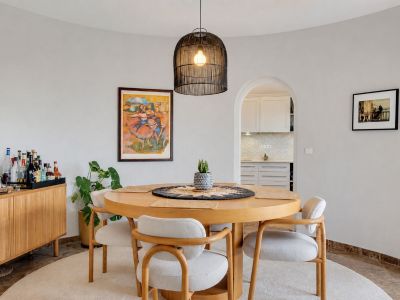 Comedor curvo con mesa redonda de madera y cuatro sillas tapizadas, iluminado por una lámpara colgante de mimbre. Un arco conecta con la cocina.