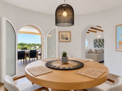 Comedor luminoso con mesa redonda de madera y lámpara de mimbre, conectado a una terraza exterior con vistas a la vegetación. Un arco conduce a la sala de estar.
