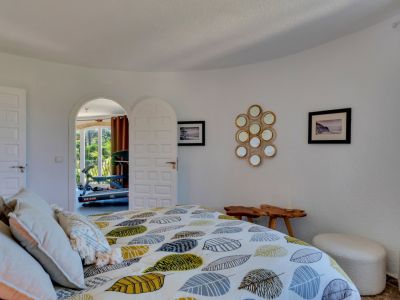 Dormitorio con paredes blancas texturizadas, cama de diseño con edredón de hojas y acceso arqueado a una sala luminosa con equipo de gimnasio.