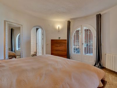 Dormitorio con puertas dobles arqueadas blancas y rejas al exterior, suelo de efecto mármol y cómoda de madera. Un gran espejo ornamental y paredes texturizadas completan la estancia.