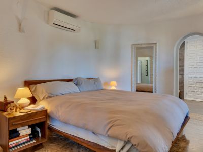 Dormitorio principal con cama de madera, paredes blancas texturizadas y aire acondicionado. Un espejo ornamentado y un arco conducen a una puerta de diseño.