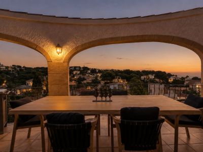 Elegante terraza con arcos al atardecer, equipada con una mesa de comedor de madera. Ofrece vistas panorámicas del área residencial iluminada y el horizonte marino.
