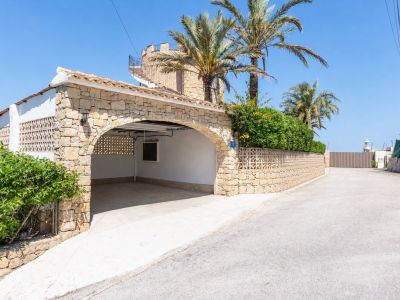 Entrada arqueada de piedra al garaje cubierto, integrada en la villa de estilo mediterráneo con escalera de caracol a la terraza superior. Palmeras y vegetación exuberante.