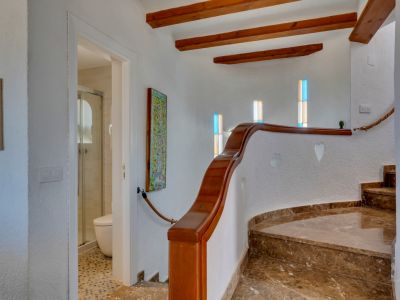Escalera curva de mármol marrón con barandilla de madera, vigas expuestas y ventanas con vidrios de colores. Acceso a un baño con ducha.